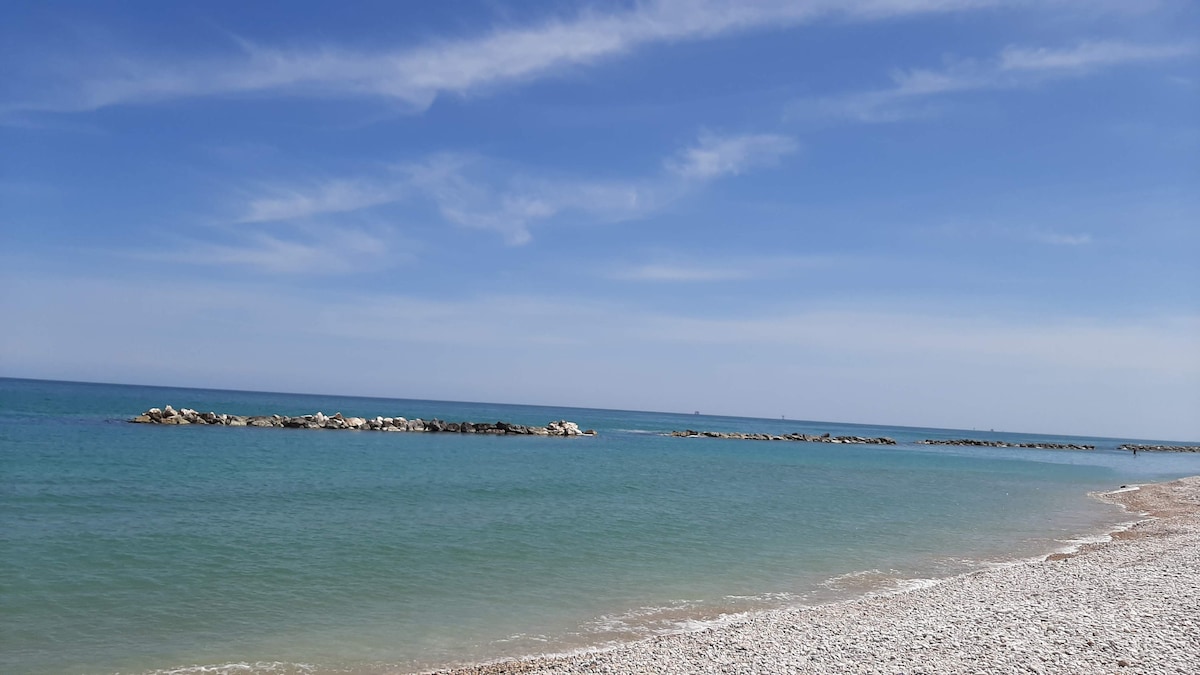 A tranquil beach scene is captured, featuring calm, clear waters meeting a pebbled shore. Smooth rocks are visible in the distance, creating a gentle contrast with the blue sky dotted with soft clouds.