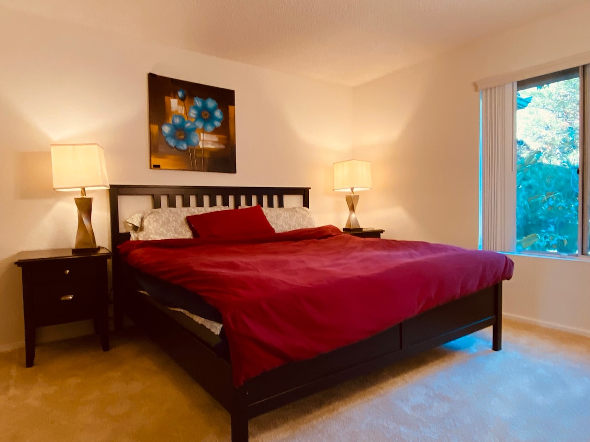 A spacious sleeping area features a large bed with a deep red comforter, framed by two bedside tables. Two lamps provide soft lighting, while a piece of wall art adds a touch of color. A window allows natural light to illuminate the room.