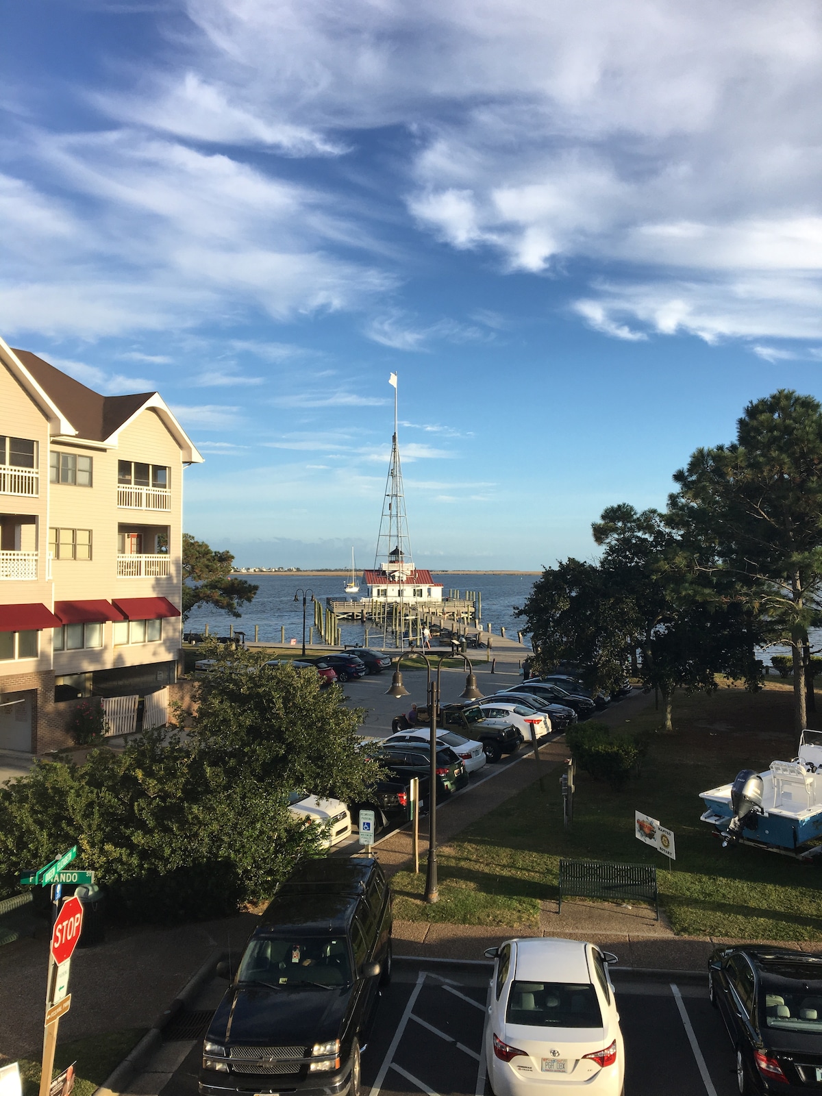 A view of Shallowbag Bay is captured from the second-floor apartment, showcasing a dock with a sailboat. Nearby buildings and parked cars are visible along the waterfront, framed by green trees under a blue sky with scattered clouds.