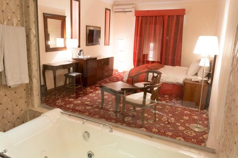 A spacious hotel room is visible through a large glass window. A comfortable bed is placed against a wall adorned with warm, neutral tones. A seating area features two chairs and a wooden table, while modern amenities include a flat-screen TV and a minibar.