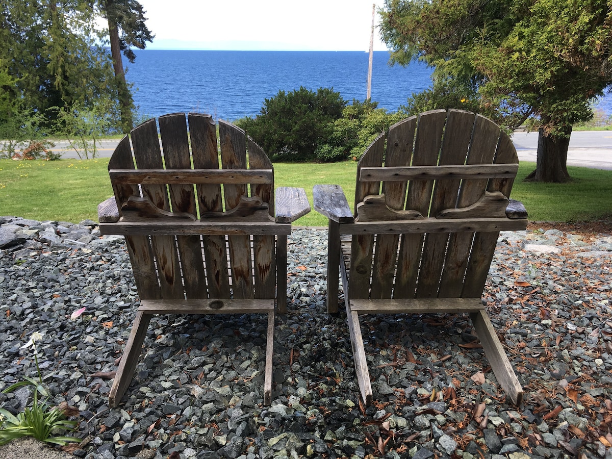 Two weathered wooden adirondack chairs are positioned facing the ocean, offering a view of the water in the distance. Surrounding the chairs is a gravel area, complemented by greenery and trees that frame the scene.