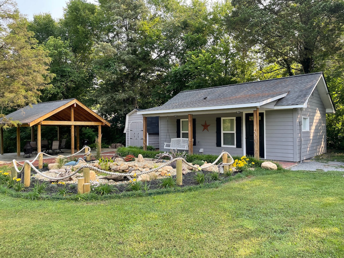 The exterior of a charming converted house is depicted, surrounded by lush greenery. A covered pavilion is visible to the left, featuring a seating area. A lovely rock garden is situated in front of the house, creating an inviting outdoor space.