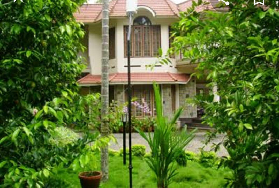 A charming homestay is surrounded by lush green fruit trees and foliage. The two-story building features a welcoming façade with large windows and a tiled roof. A well-maintained garden is visible in the foreground, complemented by potted plants and vibrant greenery.