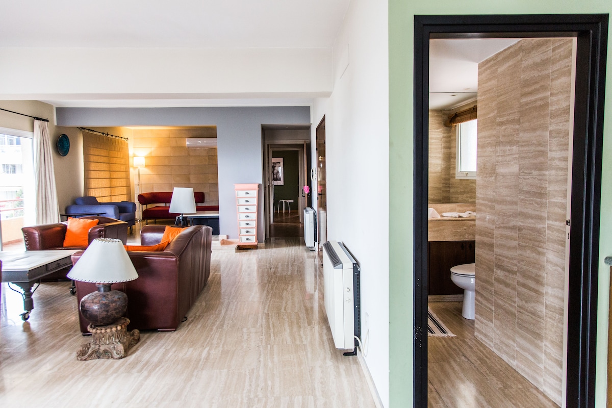 A spacious living area is viewed from the entrance, featuring a mix of bright and neutral colored furnishings. A large coffee table is placed in front of a comfortable sofa. An open doorway reveals a bathroom with tiled walls and visible fixtures, ensuring a bright and airy feel.