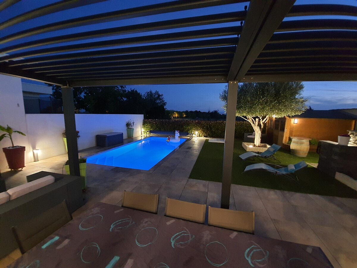 A well-lit outdoor space is showcased featuring a pool illuminated in a calming blue hue. Comfortable loungers and a dining table are arranged under a slatted pergola. Lush greenery surrounds the area, offering a serene atmosphere suitable for relaxation.