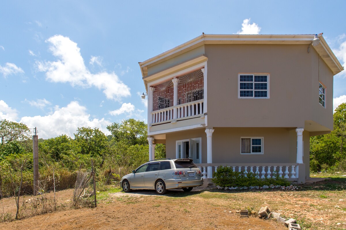 A two-story house is set against a clear blue sky, surrounded by lush greenery. A vehicle is parked nearby on the spacious dirt driveway. The house features a large balcony with decorative iron railings, emphasizing its welcoming façade.