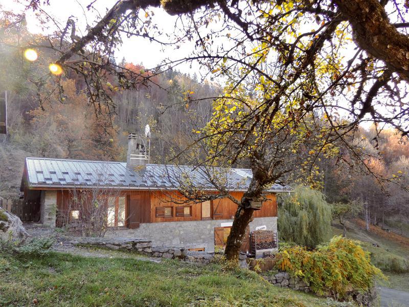 The chalet is nestled among trees, showcasing a wooden exterior complemented by stone detailing. The sloped roof is crafted from metal, reflecting the natural tones of the surrounding landscape. Autumn foliage enhances the serene setting, providing a welcoming and peaceful ambiance.