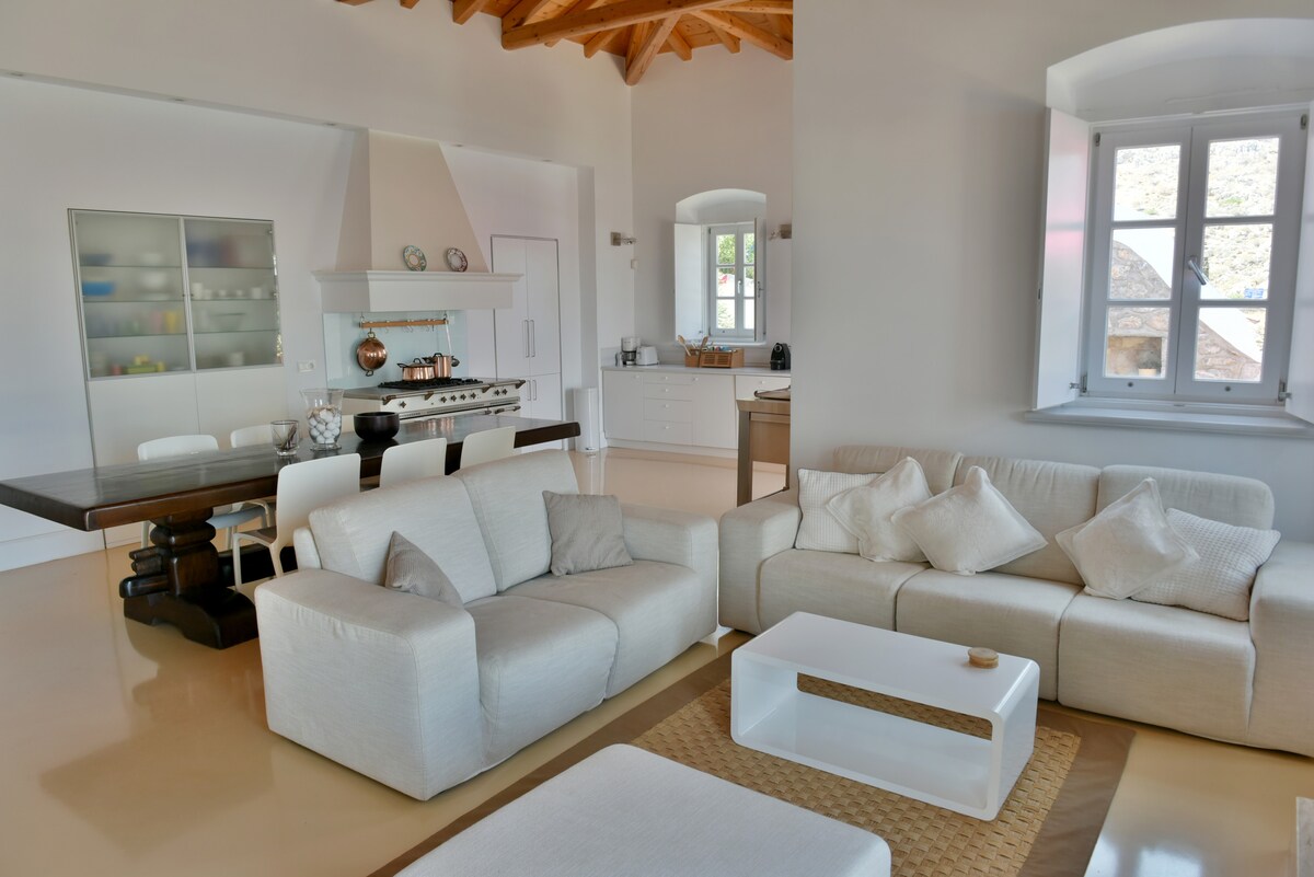 The spacious living area features a modern kitchen with sleek cabinetry and a dining table set for six. Comfortable white sofas are arranged around a minimalist coffee table, while natural light streams through large windows, highlighting a warm color palette and high wooden ceilings.