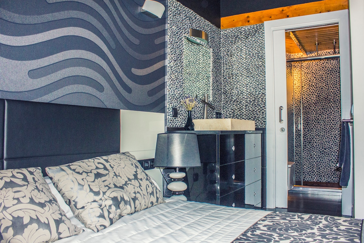 A modern bedroom features a stylish bed with decorative bedding and a bedside lamp. A sleek dresser is positioned nearby. In the background, a bathroom with a walk-in shower is visible through a glass door, displaying contemporary tile work and fixtures.