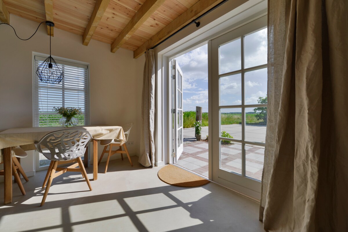 A bright dining area is shown with a wooden table and stylish chairs. Large glass doors open to the outdoor space, allowing natural light to fill the room. Decorative curtains frame the doorway, and a small rug is placed at the entrance.