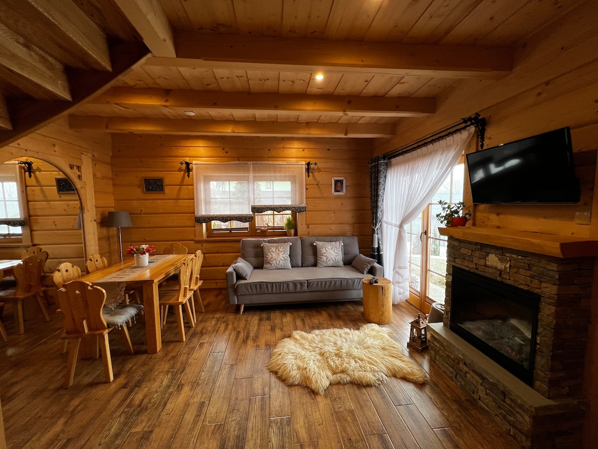 The interior features wooden paneling and a warm atmosphere. A cozy gray sofa is placed near a stone fireplace, with a light rug positioned on the wooden floor. A wooden dining table with chairs is visible, complemented by natural light from large windows.