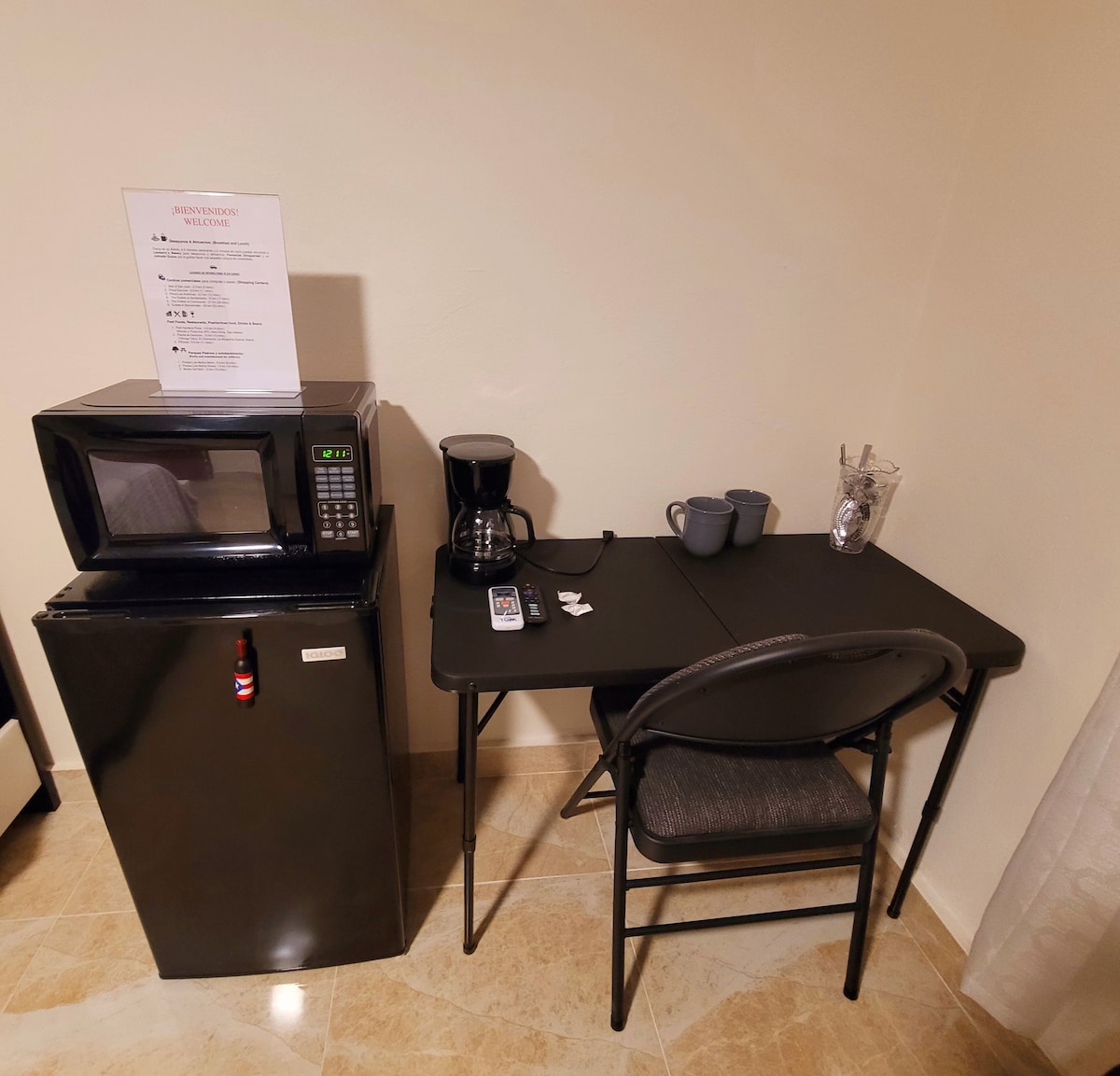 A compact kitchenette area features a black microwave positioned above a small refrigerator. Next to it, a simple black table is complemented by a chair. A coffee maker sits on the table, along with two mugs and a clear container holding utensils.