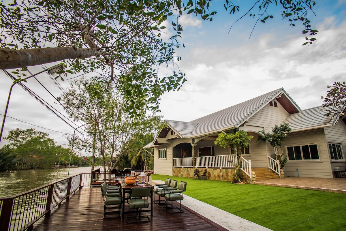 The exterior of the house is visible, featuring a spacious wooden deck surrounded by a well-maintained lawn. Green outdoor seating is arranged around a large dining table, providing a relaxing view of the nearby water. Trees and greenery enhance the natural setting.