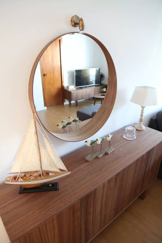 A wooden sideboard is featured with a decorative model sailboat displayed prominently. A round mirror reflects the space, enhancing depth. A lamp and small glass piece add charm, while a television is visible in the background, contributing to the room's inviting ambiance.