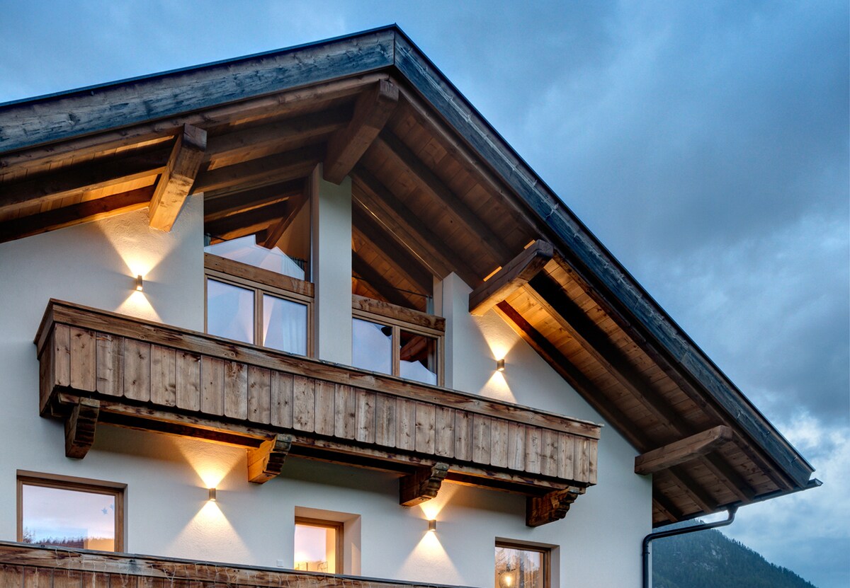 The exterior of the house features a traditional alpine design with wooden accents and a sloped roof. Warm light is emitted from wall-mounted fixtures, highlighting the balcony. Five windows offer views of the surroundings, while a serene sky creates a peaceful backdrop.
