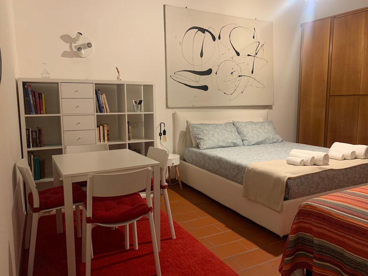 A well-lit room features a spacious bed adorned with towels. A modern artwork hangs on the wall above the bed. A small table with chairs and a bookshelf create a functional space, while warm-colored tiles cover the floor.