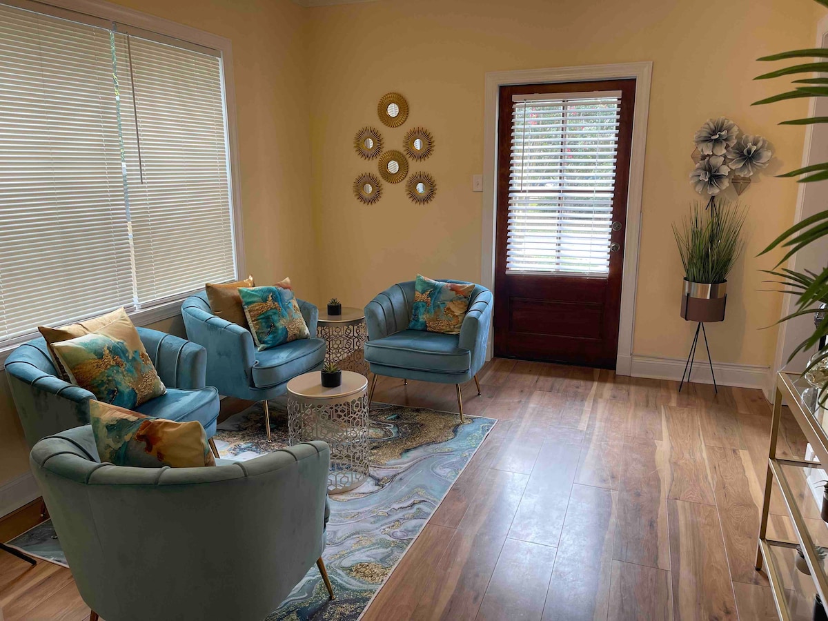 A cozy seating area is presented with two light blue chairs and a matching chair, all adorned with decorative pillows. A small, circular table sits at the center, and a large area rug enhances the space. Natural light enters through nearby windows.