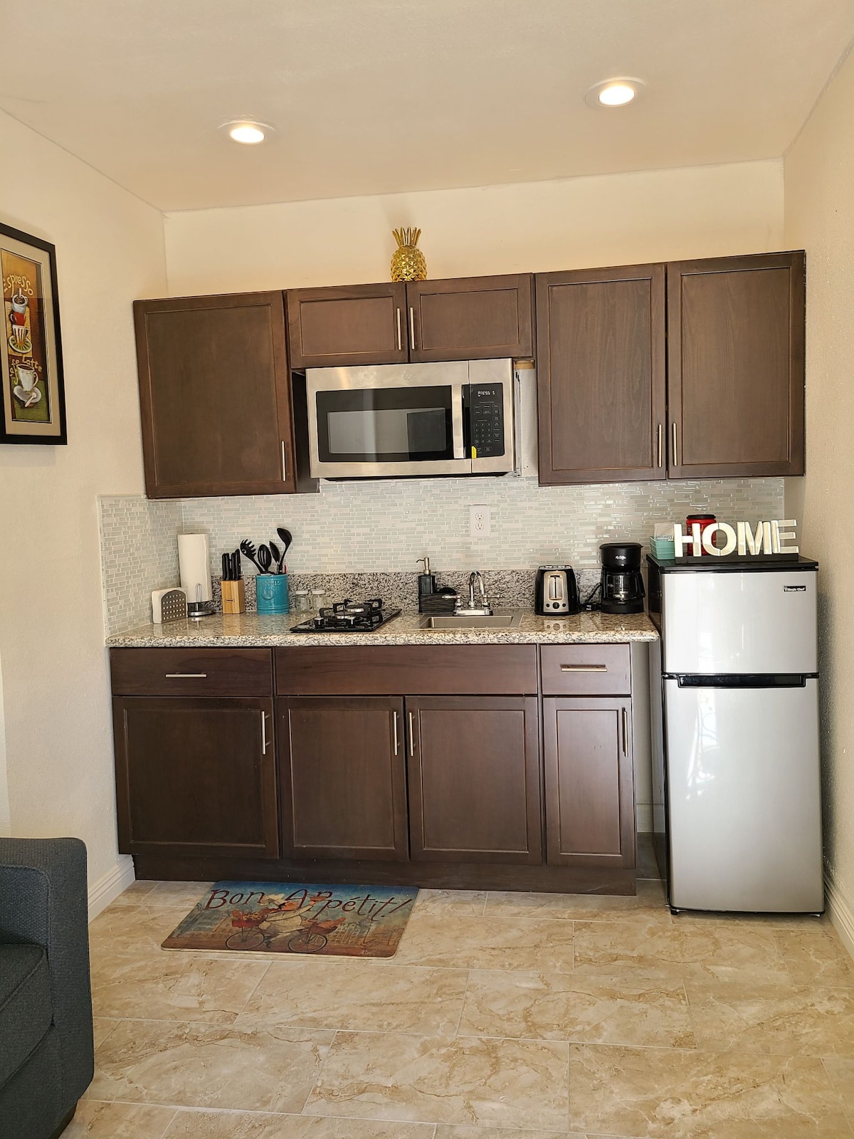 A compact kitchenette is shown, featuring dark wood cabinets and a countertop with a smooth surface. A full-size refrigerator and microwave are visible, along with a coffee maker and cooking utensils arranged neatly. A decorative rug adds a touch of hominess to the space.