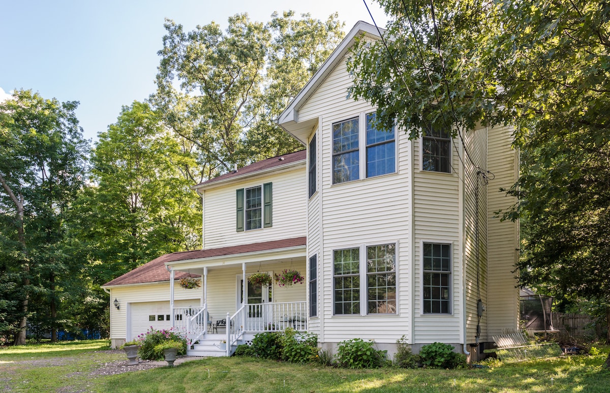 The exterior of a two-story home is framed by lush greenery, showcasing a covered front porch with hanging flower planters. Large windows allow natural light to fill the interior, while a well-kept yard complements the inviting atmosphere of the property.