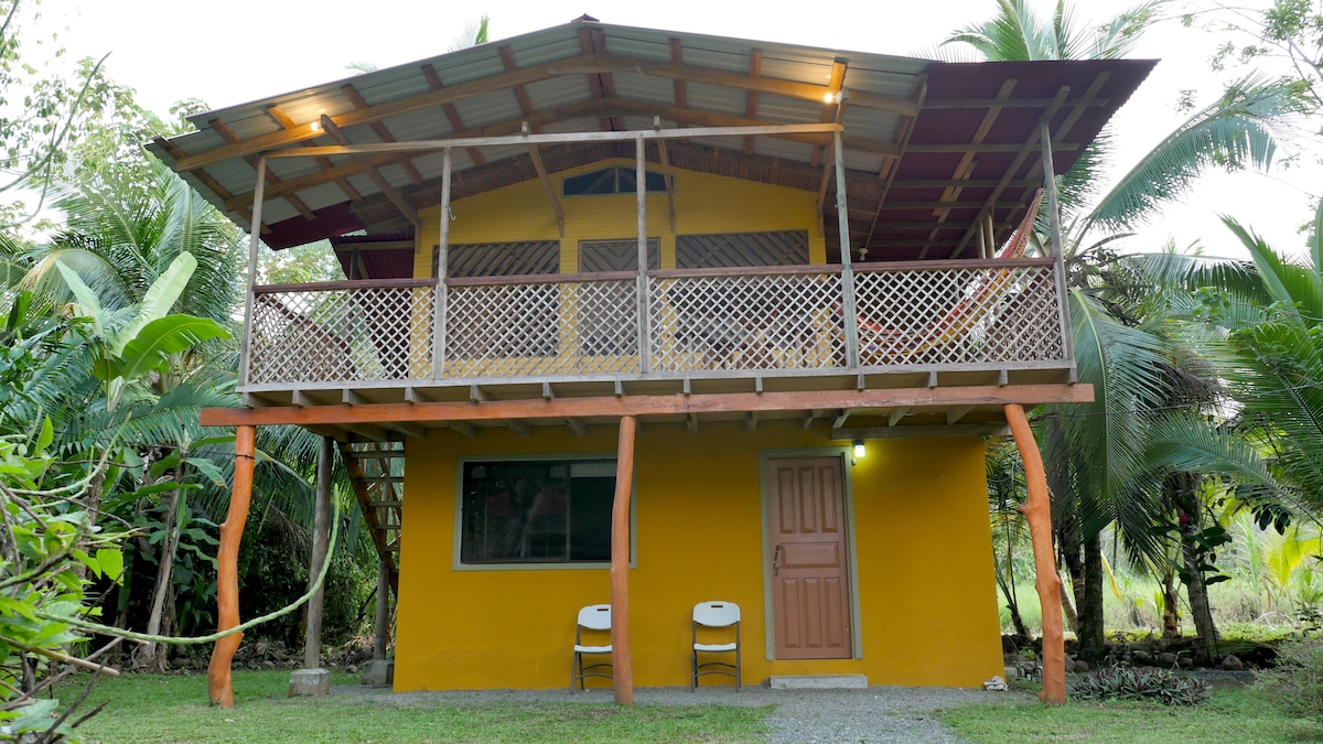 The two-story house features a vibrant yellow exterior. A spacious balcony is supported by wooden posts and is enclosed with decorative lattice. Two white chairs are placed at the entrance, while lush greenery surrounds the property, creating a tranquil setting.