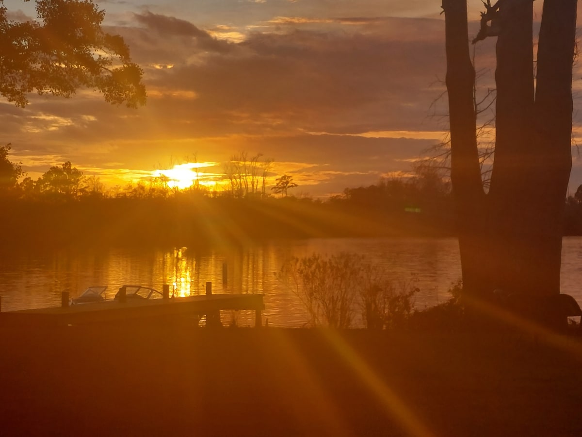 A serene sunset is depicted over the water, with golden rays reflecting on the surface. Silhouettes of trees frame the view, while a dock can be seen in the foreground. The calm atmosphere invites relaxation and connection with nature.