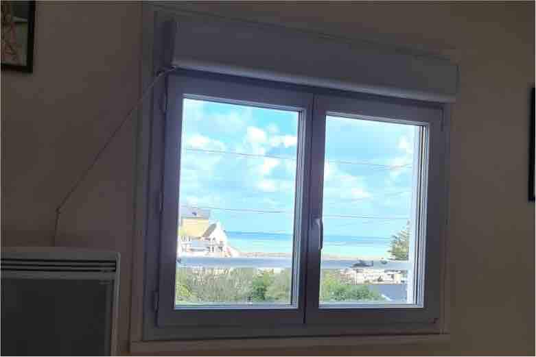 A window offers a view of the sea and nearby houses. Soft clouds are visible in the blue sky, while greenery is seen in the foreground, enhancing the connection to the surrounding landscape.