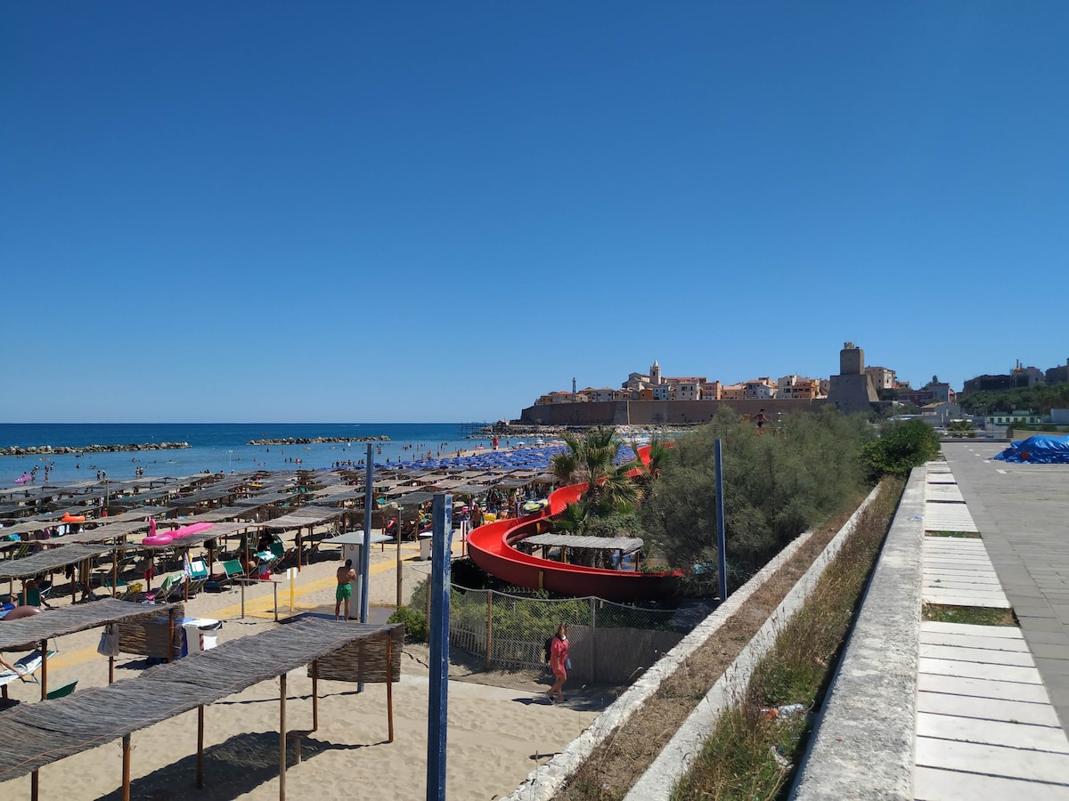 A sandy beach scene reveals numerous sun loungers and umbrellas arranged neatly along the shore. The vibrant blue sea stretches out towards the horizon, with a picturesque cliffside town visible in the background. A colorful boat is docked nearby, adding to the lively coastal ambiance.