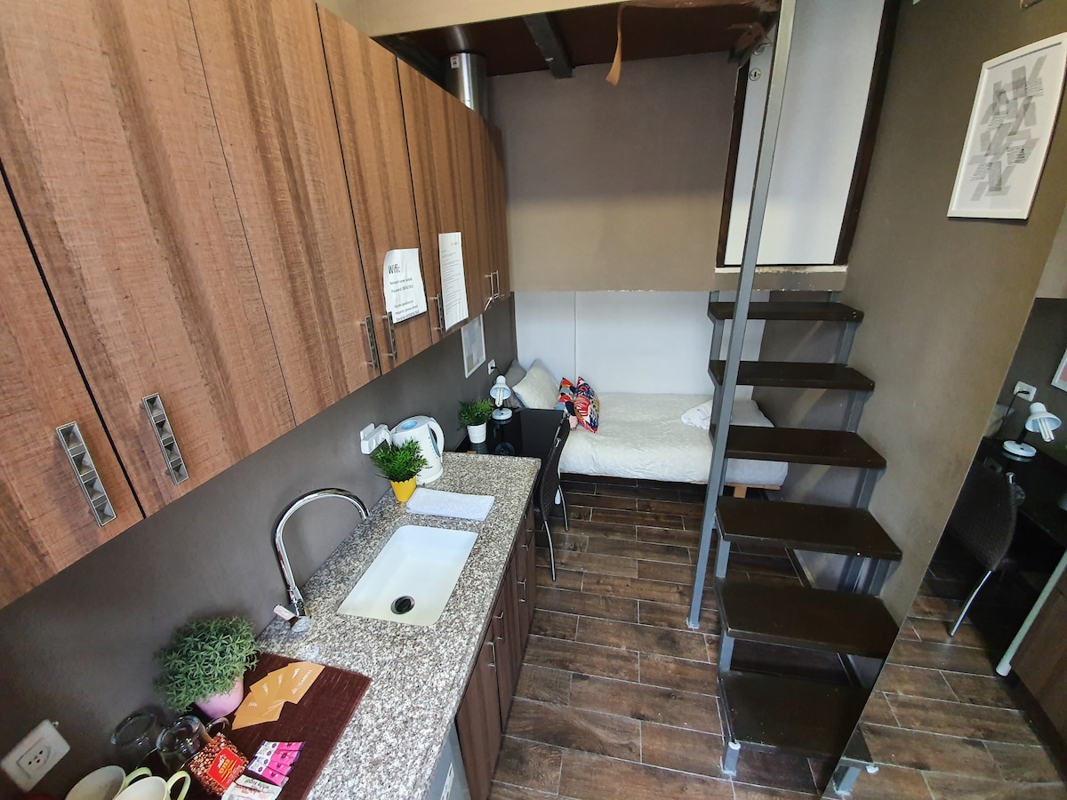 The image displays a compact studio kitchenette with wooden cabinets, a sink, and a countertop. A small dining area can be observed near the bed, which is partially visible on the upper level. A staircase leads to the sleeping area, showcasing the dual-level layout.