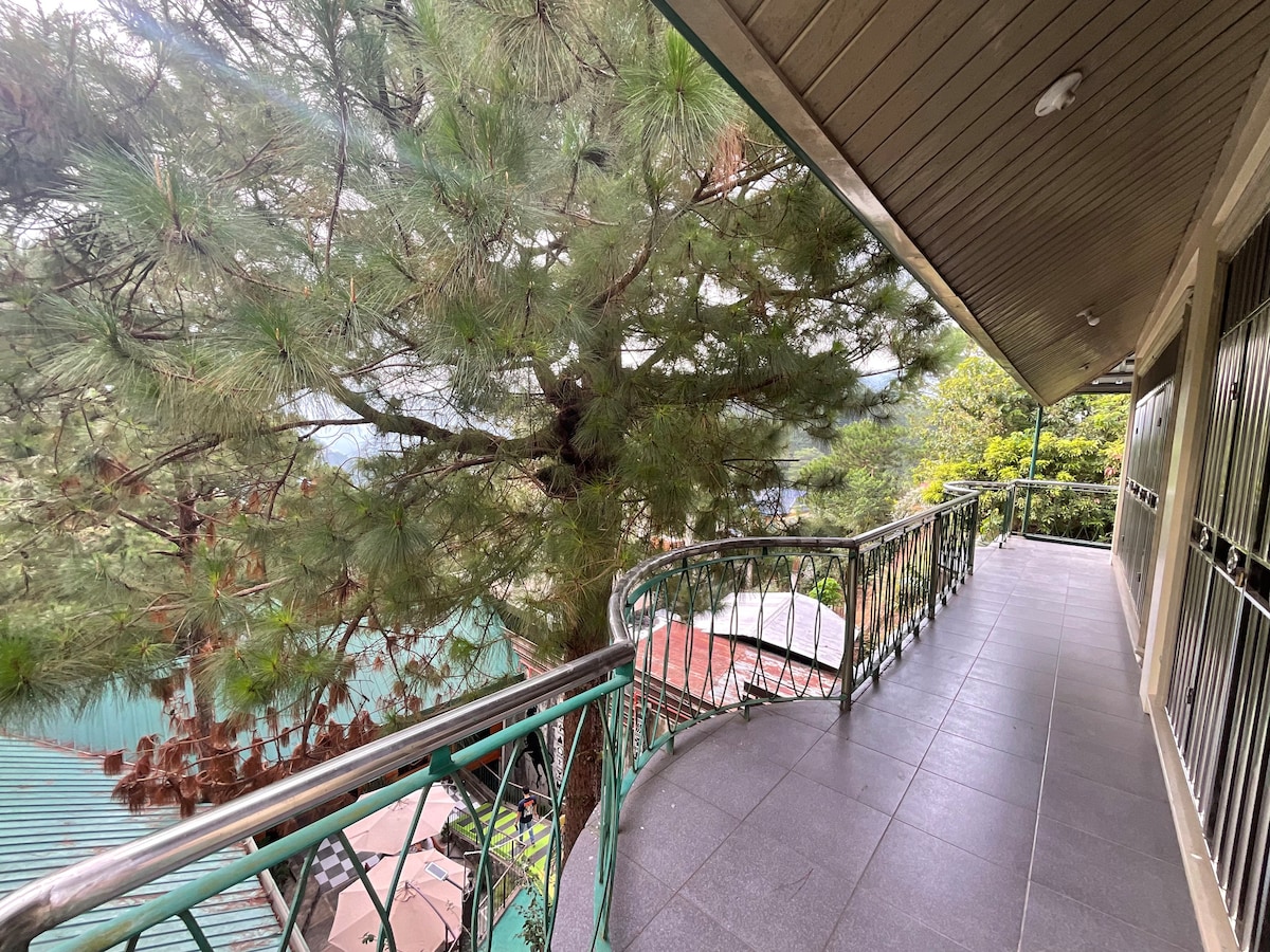 A balcony with a curved railing is shown, surrounded by tall pine trees. The floor is covered in dark tiles, and a serene view of the mountains and nearby rooftops can be seen in the distance. The setting reflects a peaceful outdoor space.