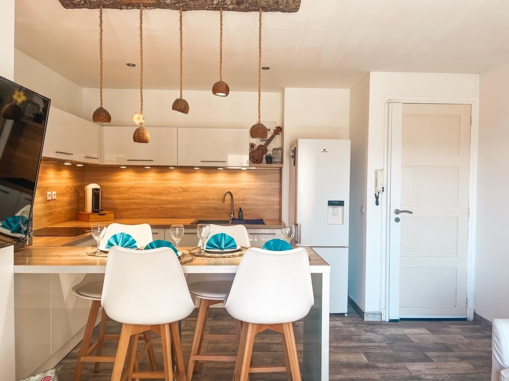 A modern kitchen area is highlighted by a wooden countertop and white cabinetry. Three white chairs are positioned at the counter, with bright blue dishware placed on the table. Warm lighting enhances the inviting atmosphere, complemented by natural wood elements overhead.