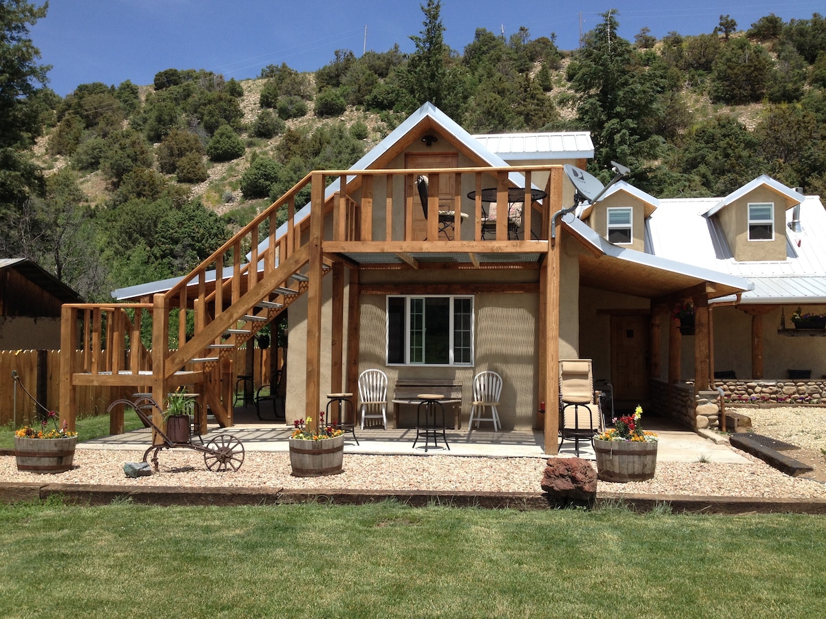 The exterior of the home is showcased with a spacious patio, wooden steps leading to a balcony, and two chairs positioned at a small table. Surrounding flower planters and well-maintained lawn areas add to the inviting atmosphere against a backdrop of mountains and greenery.