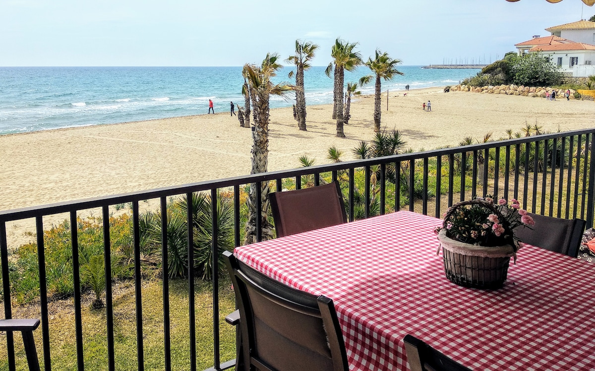 A balcony table set for dining is surrounded by sturdy chairs, positioned to offer unobstructed views of the sandy beach and ocean. Lush palm trees and shrubbery frame the scene, while a floral centerpiece adds a touch of nature to the outdoor dining space.