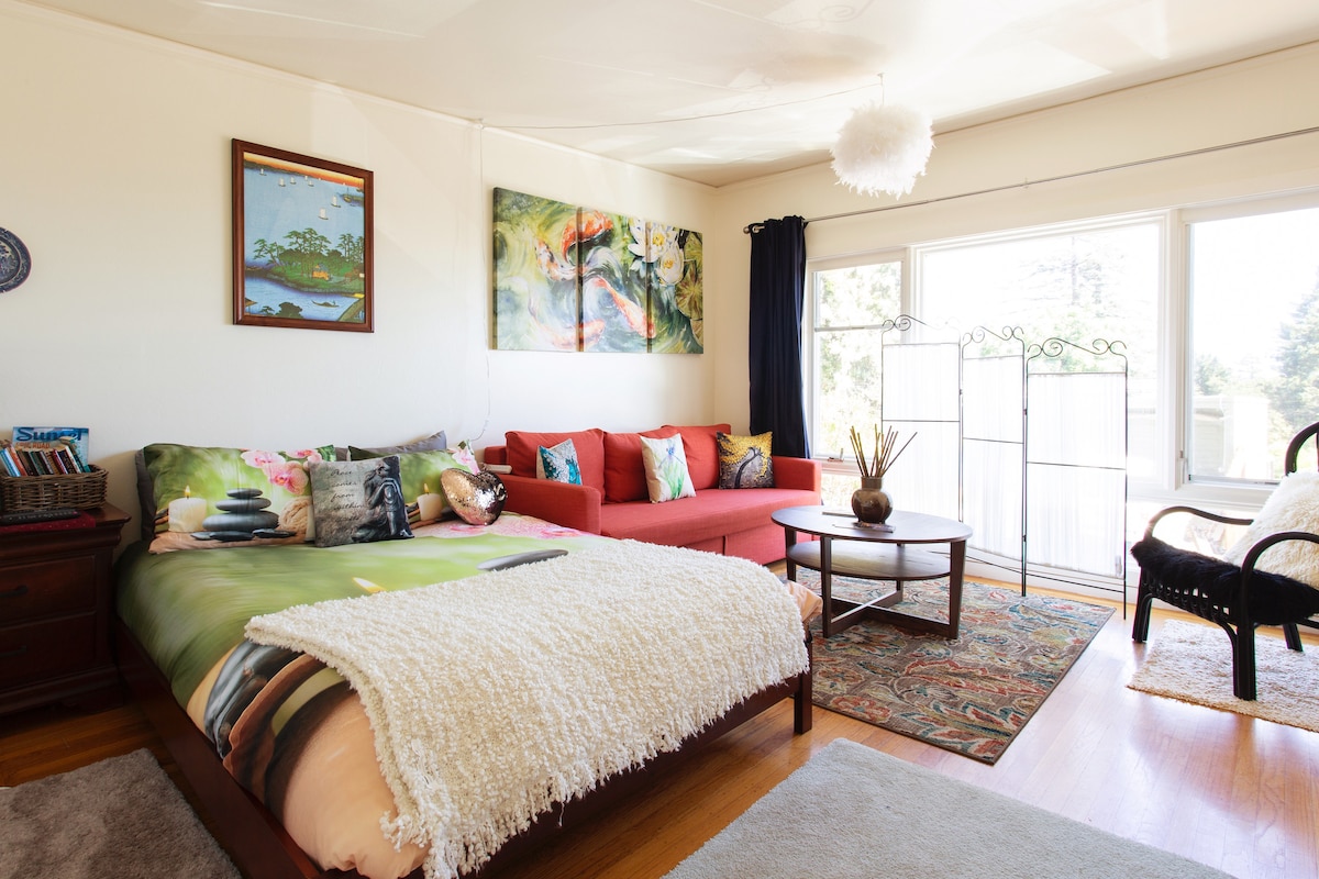A bright studio space features a spacious bed adorned with a colorful comforter and decorative pillows. A cozy seating area contains a red sofa and a wooden coffee table, with a patterned rug underneath. Large windows provide ample natural light and showcase outdoor views.