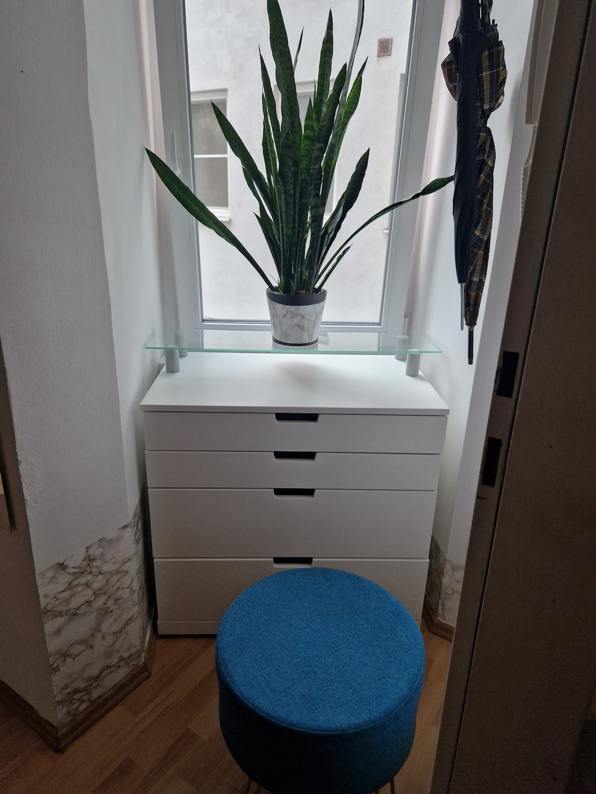 A modern storage unit featuring six drawers is positioned near a large window. A green potted plant is placed on top, complemented by a clear glass shelf. A round, blue stool is situated nearby, providing a cozy seating option.