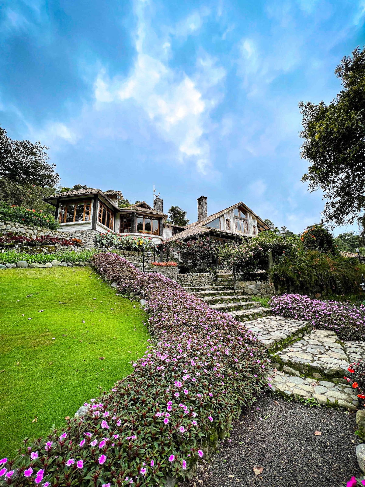 A charming house is set against a backdrop of a cloudy sky, surrounded by lush greenery. A stone pathway is lined with vibrant flowers, leading to the entrance. The architecture features large windows that enhance the connection with the surrounding nature.