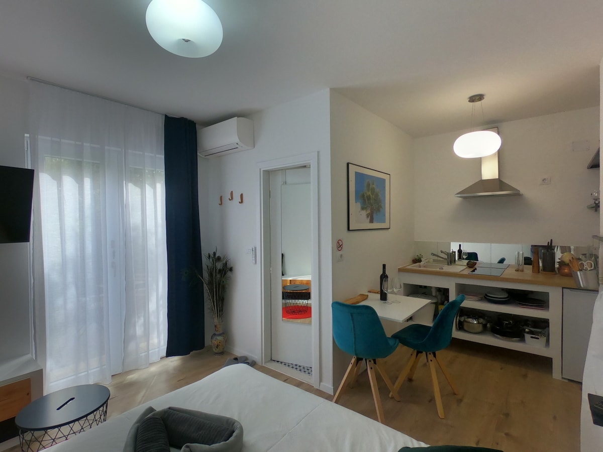 An open space studio apartment features a compact kitchen area, dining table with blue chairs, and a comfortable sleeping area. Large windows allow natural light, complemented by neutral decor and minimalistic furnishings. Air conditioning and a Smart TV are included for convenience.