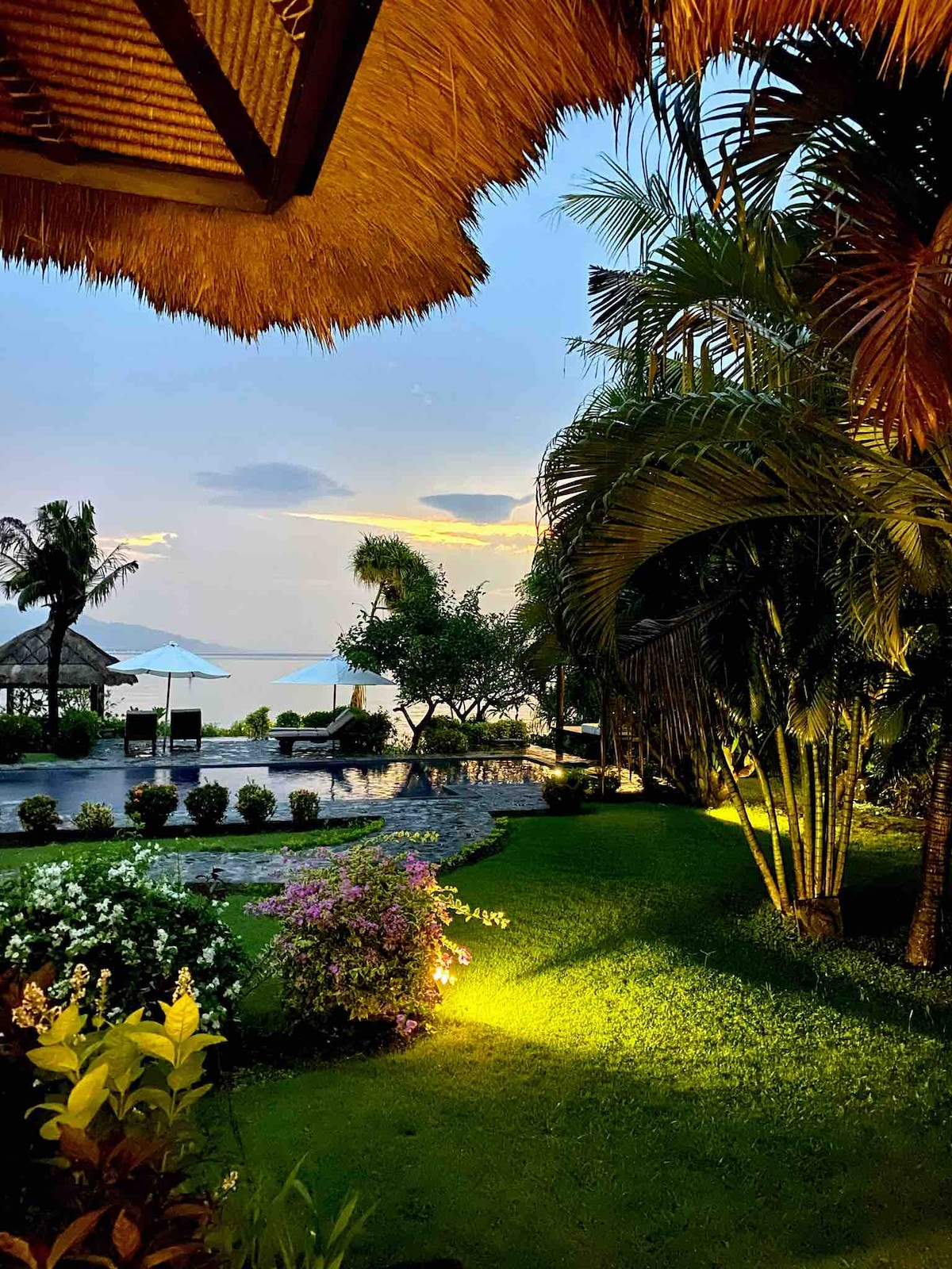 The image captures a lush garden area illuminated by soft lighting, featuring vibrant greenery and blooming flowers. In the background, a private pool reflects the tranquil sea with palm trees framed against a colorful sunset sky.