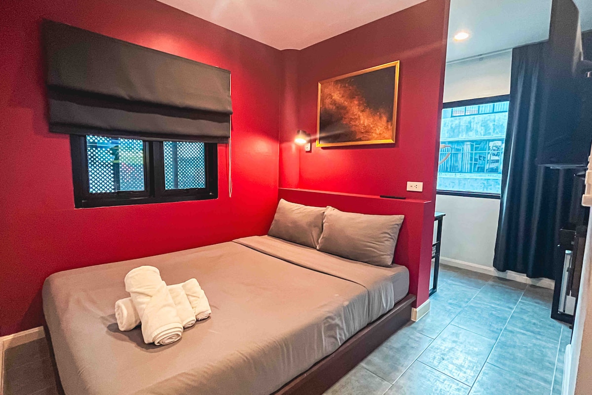 A cozy sleeping area is highlighted by deep red walls. A spacious bed is neatly made with gray linens, featuring fresh white towels placed on top. Natural light enters through a window, which is covered by a sleek black blind, creating a comfortable ambiance.