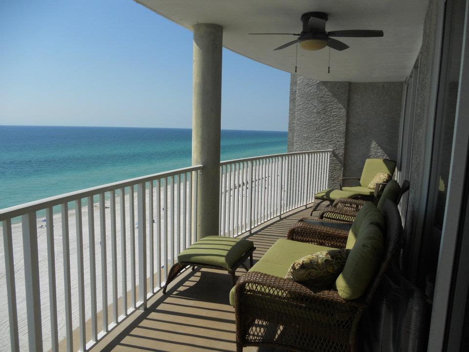 A spacious oceanfront balcony is furnished with multiple lounge chairs. The railing provides an unobstructed view of the beach and turquoise waters. A ceiling fan is visible above, complementing the serene coastal environment.