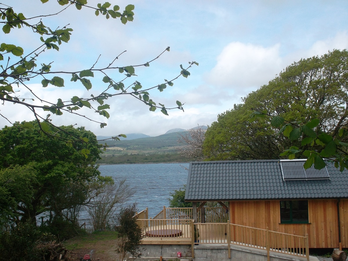 A wooden cabin stands alongside Kenmare Bay, framed by lush greenery and gentle hills. A spacious deck provides outdoor seating areas, with a glimpse of a hot tub. The serene water reflects the cloudy sky, creating a peaceful atmosphere.
