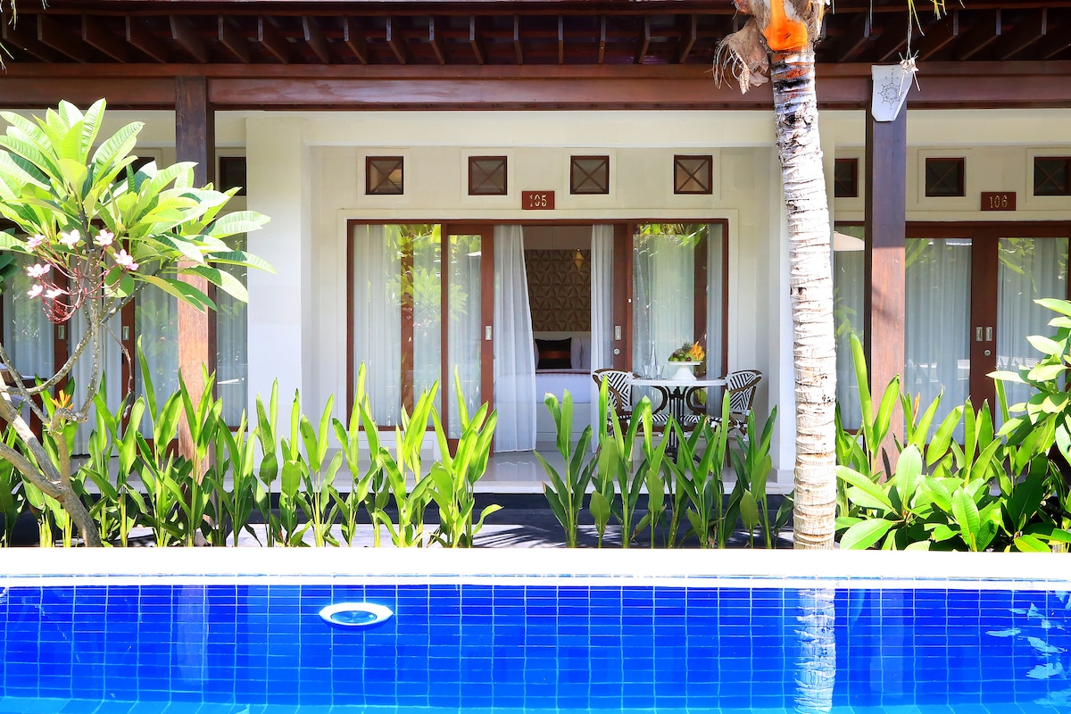 A vibrant green courtyard surrounds a serene swimming pool, with lounge chairs visible under the shade of palm trees. Large glass doors reveal a welcoming seating area, framed by tropical plants, creating a peaceful atmosphere for relaxation.