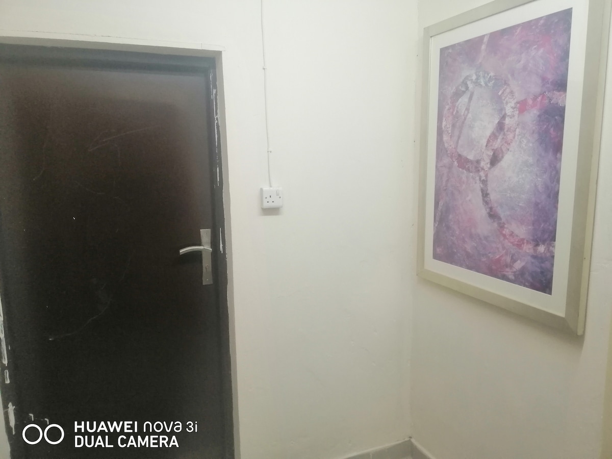 A corridor is depicted, featuring a dark door with a silver handle on the left side. A large framed art piece in shades of purple and pink is displayed on the right wall. The flooring is tiled, contributing to a clean and simple environment.