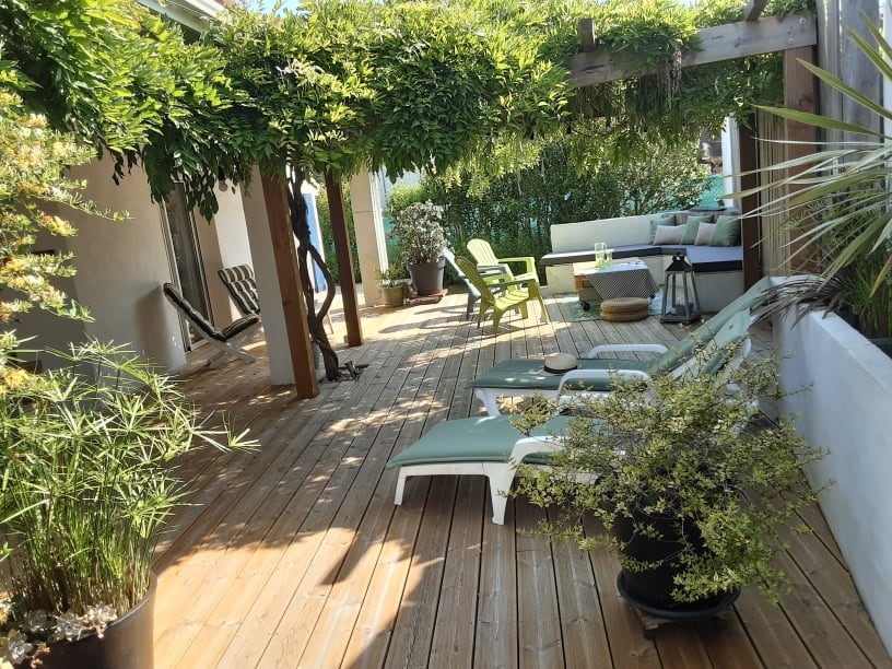 A spacious wooden terrace is featured, shaded by lush greenery and a wooden pergola. Comfortable lounge chairs are arranged for relaxation, accompanied by potted plants and a seating area with cushions. The scene reflects a serene outdoor space, ideal for enjoying time in nature.