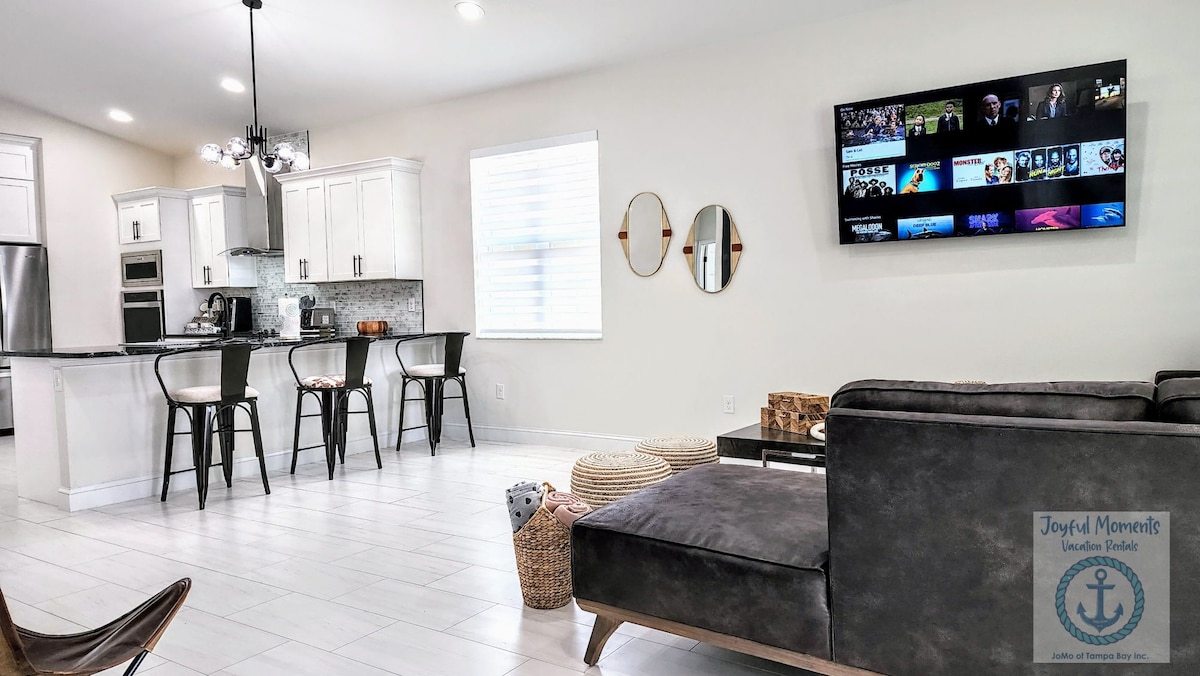 An open living space features a modern kitchen with stainless steel appliances and a breakfast bar with three high-backed chairs. A flat-screen TV is mounted on the wall, and natural light filters through the window, creating a bright and inviting atmosphere.