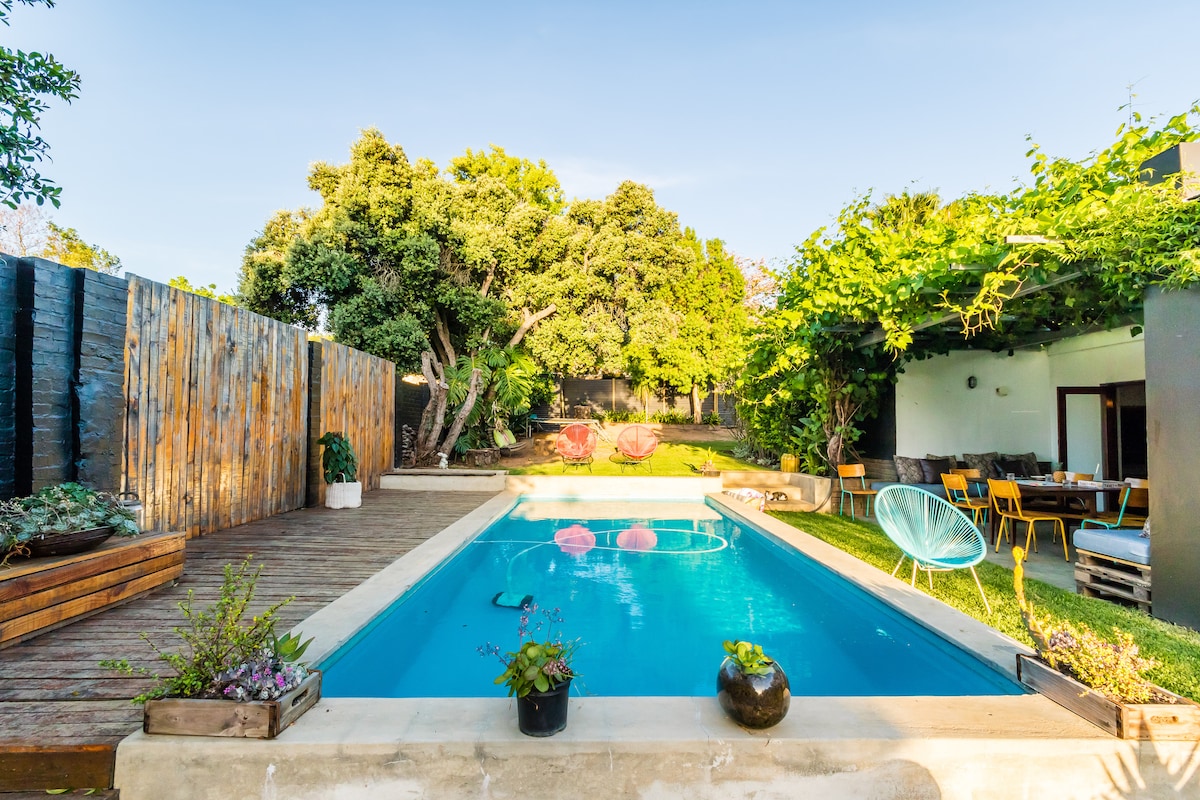 A serene swimming pool is surrounded by lush greenery and wooden decking. The pool area is complemented by vibrant planters and a comfortable seating area nearby, offering a place for relaxation. Natural light enhances the inviting atmosphere of the outdoor space.