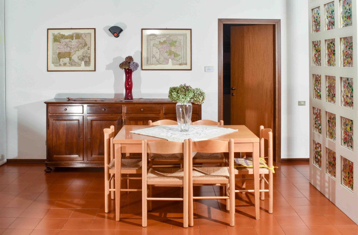 A dining area features a light wooden table surrounded by six chairs. A red vase with dried flowers is placed at the center of the table. Two framed artwork pieces hang on the wall, while a wooden sideboard offers additional storage.