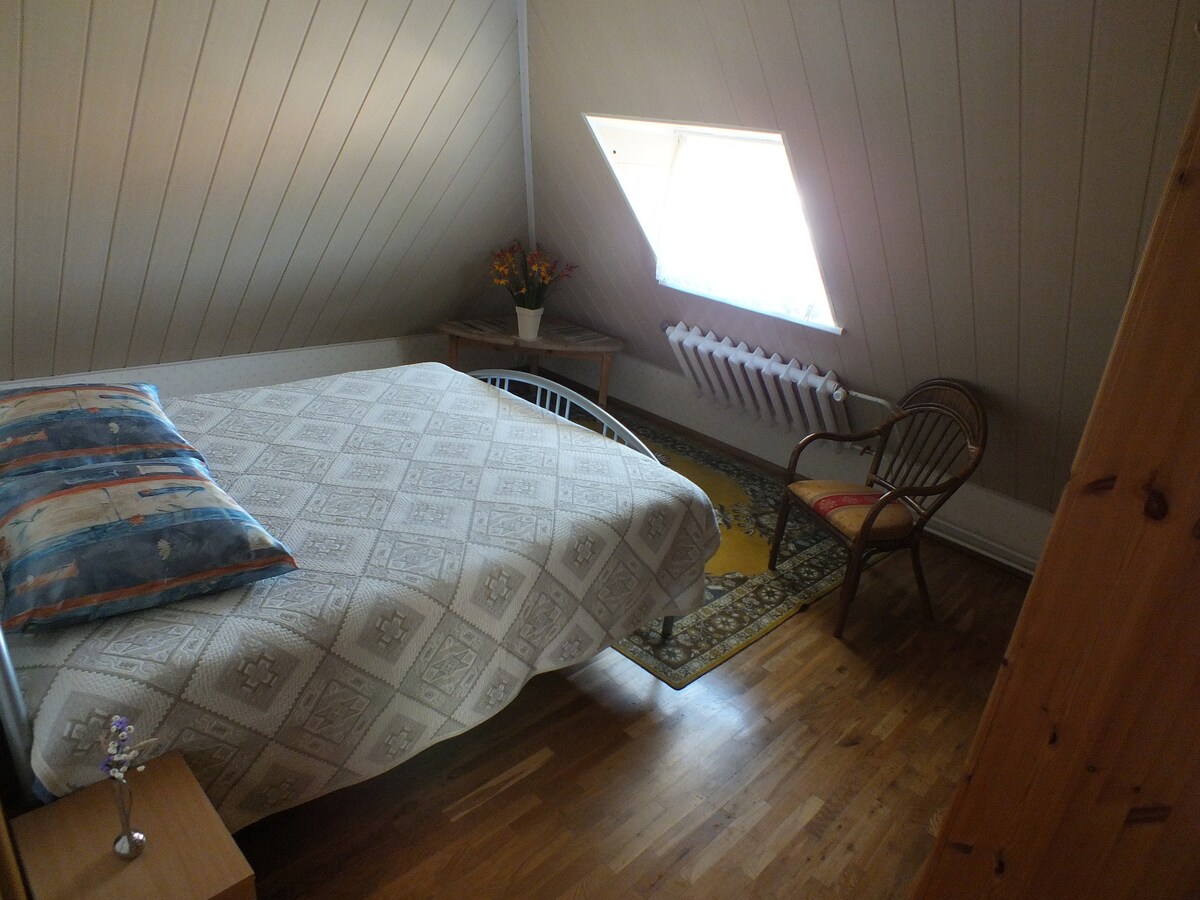 A simple family room features a spacious bed covered with a patterned blanket. A small wooden table displays a vase of flowers, and a rattan chair is positioned nearby. Natural light enters through a slanted window, illuminating the wooden floor.