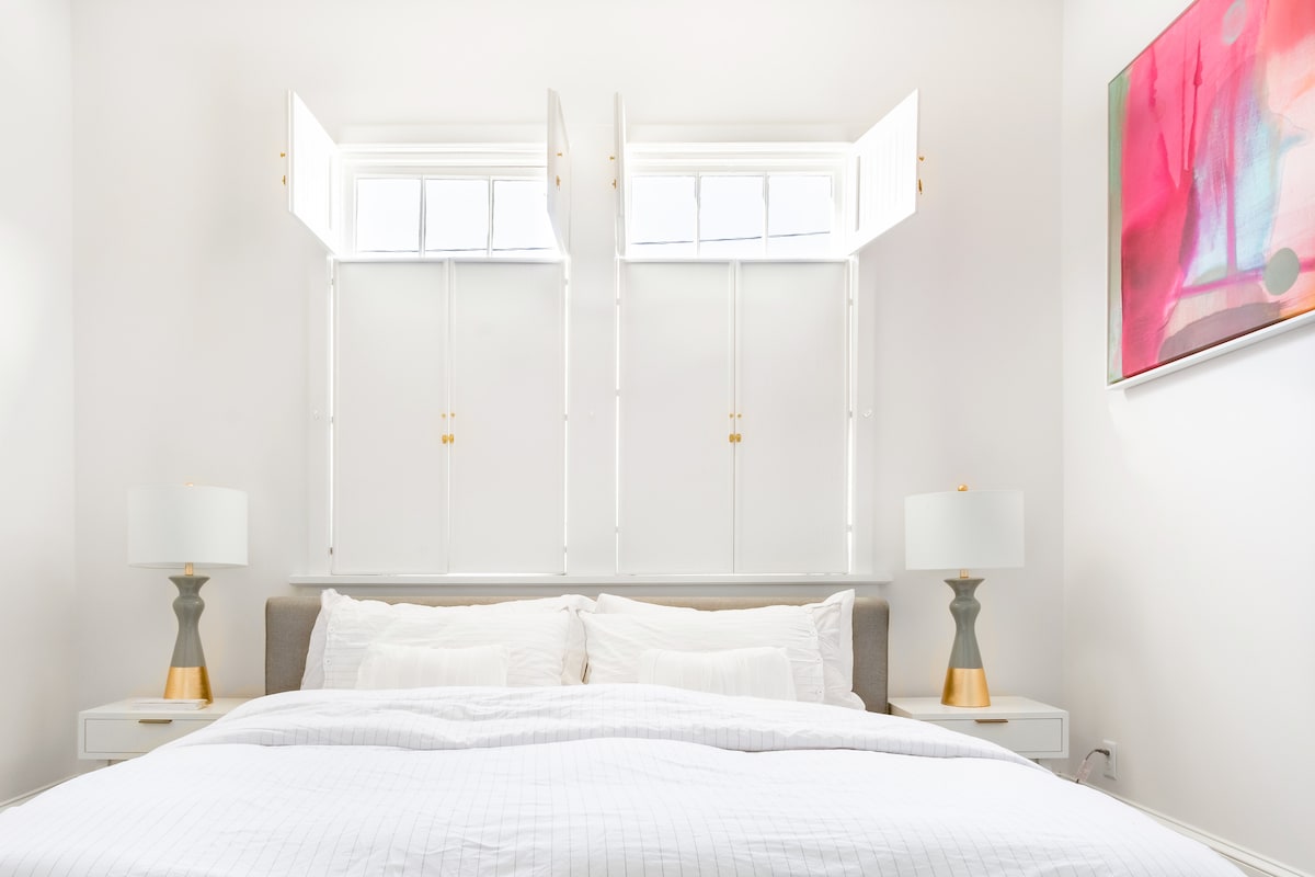 A bright and airy bedroom features a comfortable bed adorned with crisp white linens. Two windows open to let in natural light, flanked by stylish lamps on either side. A colorful piece of wall art adds a touch of vibrancy to the serene atmosphere.