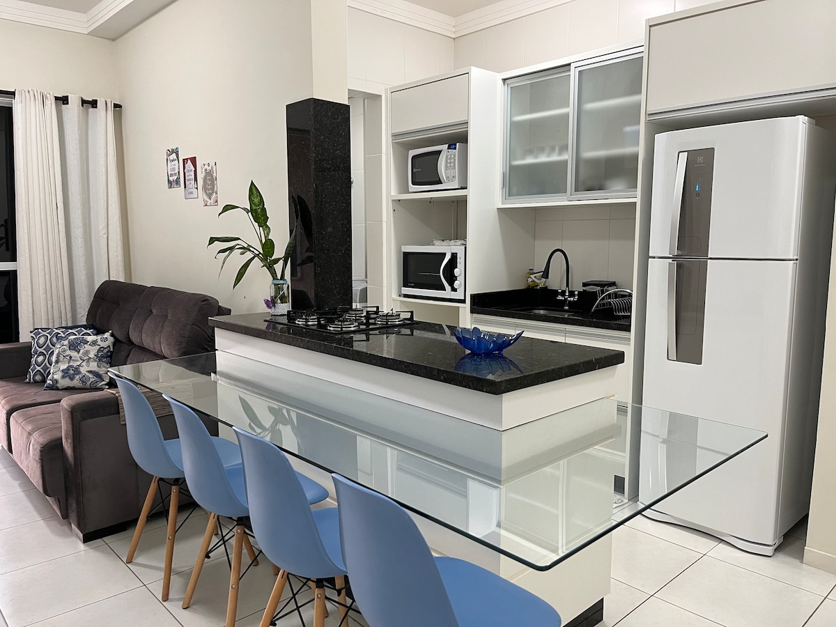 A modern kitchen and dining area features sleek cabinetry and polished surfaces. The dining table, designed with a glass top, is surrounded by several blue chairs. Appliances including a refrigerator and microwave are visible, contributing to a functional and inviting space.