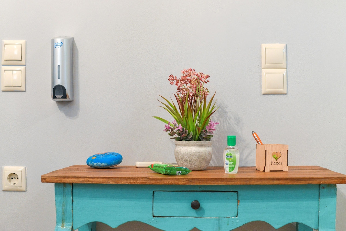 A light blue table is presented in front of a neutral wall. A small potted plant with vibrant pink flowers is displayed on the table, alongside a bottle of hand sanitizer, a wooden box, and a blue stone. Light switches and a paper towel dispenser are visible on the wall.